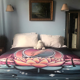 The Lotus Stealie fleece blanket on a bed with white pilllows and a stuffed bear, in front of a blue wall with a picture and two lights above the bed and a full-length mirror next to it.