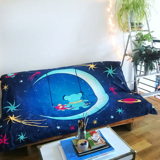 The blue Moon Swing blanket draped on a futon, with a coffee table in front of it, and a ladder with potted plants on the rungs next to it.