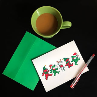 A close up aerial view of the Jingle Bears greeting card with a green envelope and a red pen, on a black surface, next to a half-filled green coffee cup